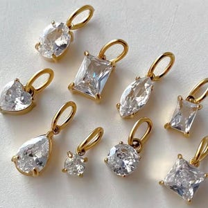 Gold diamond crystal charms displayed on a white background, each featuring a unique rhinestone cut — pear, heart, round, oval, marquise, princess, emerald, and radiant. The clear faceted stones shimmer in the light, framed in gold settings with looped bails for attaching to necklaces or bracelets. Elegant minimalist photo showing jewelry-making pendants available in both gold and silver options.  Perfect for diy jewelry to use as add-on charms for any charm necklace or charm bracelet.
