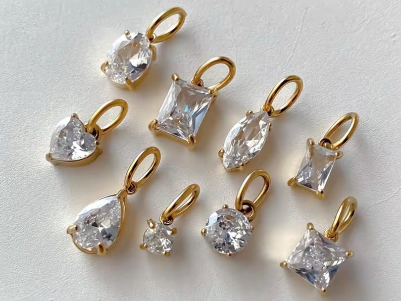 Gold diamond crystal charms displayed on a white background, each featuring a unique rhinestone cut — pear, heart, round, oval, marquise, princess, emerald, and radiant. The clear faceted stones shimmer in the light, framed in gold settings with looped bails for attaching to necklaces or bracelets. Elegant minimalist photo showing jewelry-making pendants available in both gold and silver options.  Perfect for diy jewelry to use as add-on charms for any charm necklace or charm bracelet.