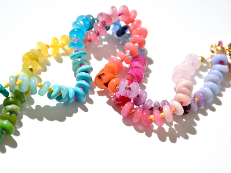 May include: A rainbow colored beaded necklace with round, flat beads. The beads are arranged in a gradient pattern, starting with green and ending with purple.