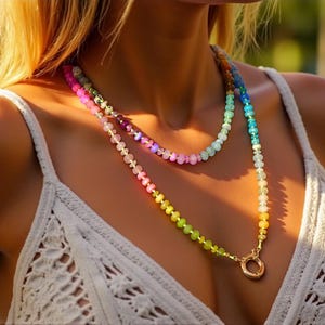 Multi-strand beaded necklace featuring a rainbow gradient of gemstone beads.  The necklace includes pink, purple, blue, green, and yellow beads, graduating smoothly from one color to the next.  The beads are faceted and appear to be semi-precious stones. The necklace has a gold clasp.  The necklace is layered, with one strand slightly shorter than the other. The style is bohemian and colorful.