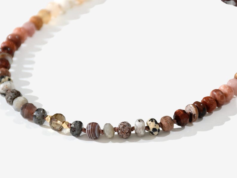 A long necklace made of various colored stones. The stones are in shades of brown, creams, white, and natural hues. The necklace has a gold clasp.