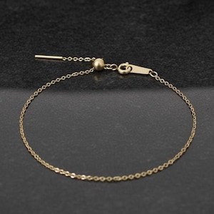 May include: Delicate gold-colored bracelet with a fine chain and adjustable clasp. The bracelet features a small, round bead and a slender bar for adjusting the length. The jewelry is displayed against a dark background.