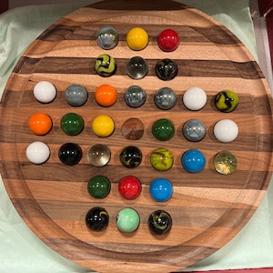 May include: A wooden game board with a circular playing surface and a pattern of alternating light and dark wood stripes. The board is filled with 25 marbles of various colors, including red, blue, green, yellow, white, and black.