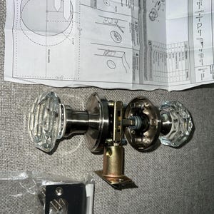 May include: A silver door knob set with clear glass knobs. The set includes a door knob, a latch, and a strike plate. The door knob is a lever style with a square shape. The glass knobs are faceted and have a crystal-like appearance.