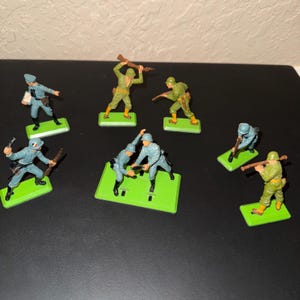 Peut inclure: Un ensemble de sept figurines de soldats en plastique dans diverses poses d'action. Les personnages sont peints en uniformes bleu clair et vert olive, chacun monté sur une base rectangulaire vert vif. Certains soldats tiennent des fusils, d'autres du matériel.