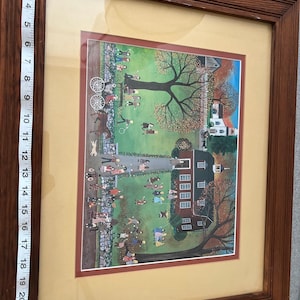 May include: Framed folk art print depicting a village scene with people, trees, and buildings. The artwork is set within a brown wooden frame, with a cream-colored mat. The print features a vibrant color palette and a detailed, whimsical style.