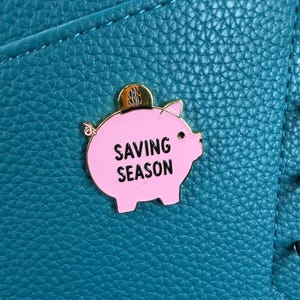 May include: A pink enamel pin shaped like a piggy bank with a gold coin slot on top. The pin features the black text SAVING SEASON and is attached to a textured teal surface. The pig has a small curly tail, a black eye, and a pink ear.