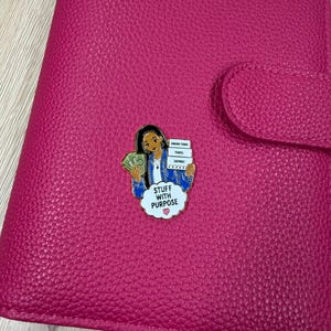 May include: A bright pink textured leather-like planner cover featuring a metal enamel pin of a person holding green paper money and a stack of white labels reading Sinking Funds, Travel, and Savings, with a speech bubble saying STUFF WITH PURPOSE.