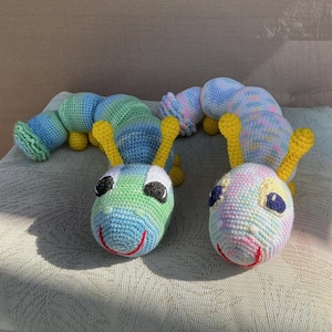 May include: Two crocheted caterpillar toys with segmented bodies. The left caterpillar features blue and green variegated yarn with yellow antennae and black eyes. The right caterpillar uses pastel multicolored yarn with yellow antennae and dark blue eyes.