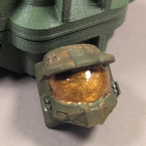 May include: A green 3D-printed box with the letters "UNSC" and a black and white emblem. A small, detailed green helmet with a gold visor is in front of the box. The box is hinged and appears to open.