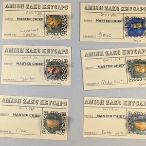 May include: Six custom keycaps featuring Master Chief helmet designs. Each keycap is mounted on a small card with the text "AMISH ZAKU KEYCAPS" and design details. Colorways include "Covenant Splatter," "Bleue," "Splatter," "Midas Dust," "Clean," and "Battle Worn."