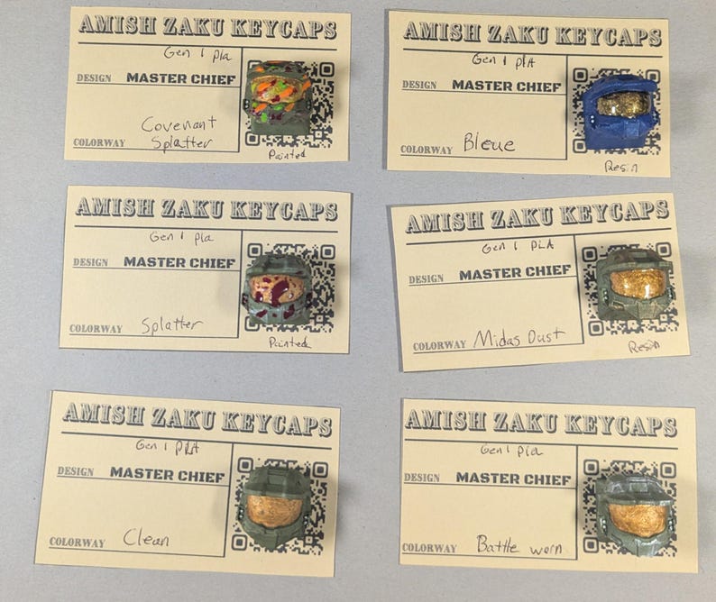 May include: Six custom keycaps featuring Master Chief helmet designs. Each keycap is mounted on a small card with the text "AMISH ZAKU KEYCAPS" and design details. Colorways include "Covenant Splatter," "Bleue," "Splatter," "Midas Dust," "Clean," and "Battle Worn."