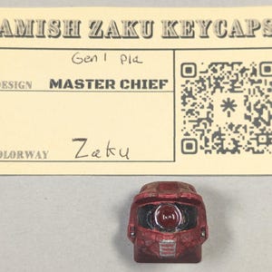 May include: A red Zaku keycap in the shape of a Master Chief helmet. The keycap is placed in front of a beige card with the text "AMISH ZAKU KEYCAPS", "MASTER CHIEF", and "Zaku". A QR code is also visible.