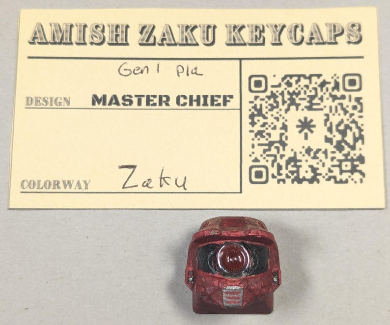 May include: A red Zaku keycap in the shape of a Master Chief helmet. The keycap is placed in front of a beige card with the text "AMISH ZAKU KEYCAPS", "MASTER CHIEF", and "Zaku". A QR code is also visible.