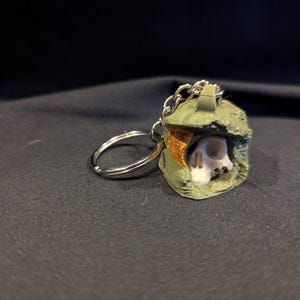 May include: A green and brown helmet-shaped keychain with a skull visible inside. The keychain has a silver chain and ring. The background is a dark, textured fabric.