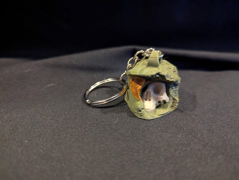 May include: A green and brown helmet-shaped keychain with a skull visible inside. The keychain has a silver chain and ring. The background is a dark, textured fabric.