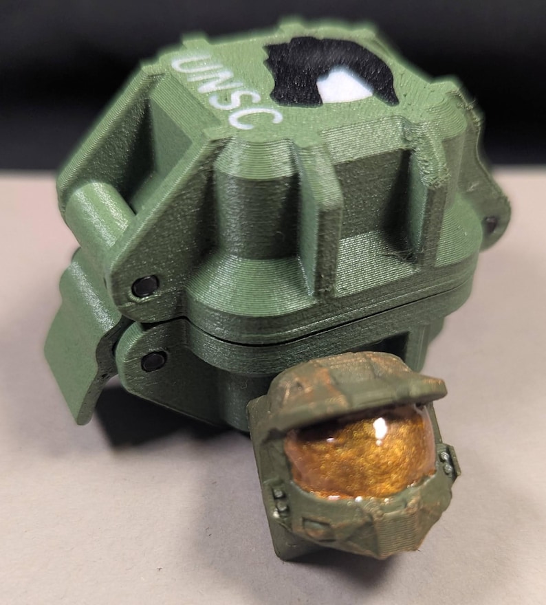 May include: A green 3D-printed box with the letters "UNSC" and a black and white emblem. A small, detailed green helmet with a gold visor is in front of the box. The box is hinged and appears to open.