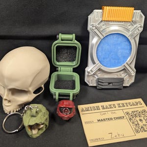 May include: An assortment of 3D-printed items, including a skull, a green hinged box, a red helmet, a silver and orange device, and a green dinosaur keychain. A card reads "Amish Zaku Keycaps, Master Chief, Zaku."