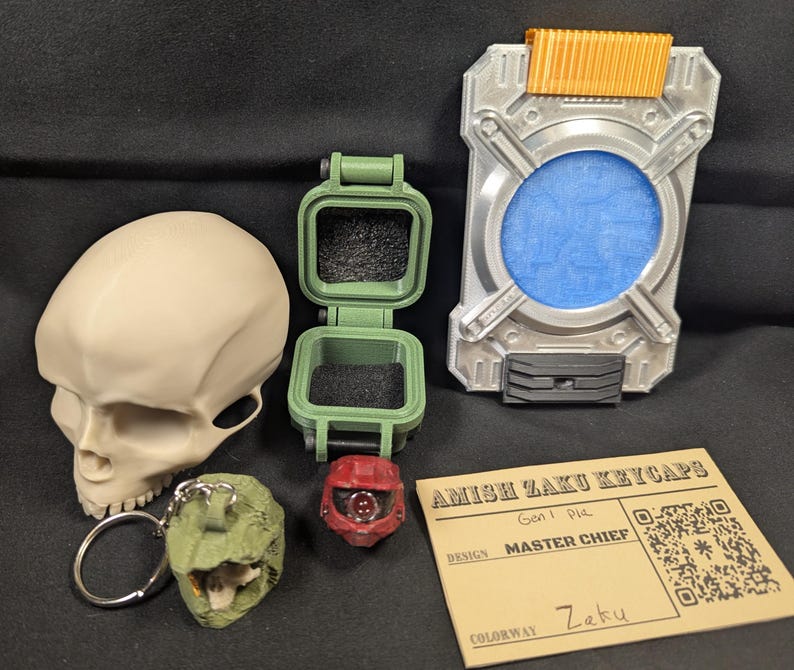 May include: An assortment of 3D-printed items, including a skull, a green hinged box, a red helmet, a silver and orange device, and a green dinosaur keychain. A card reads "Amish Zaku Keycaps, Master Chief, Zaku."