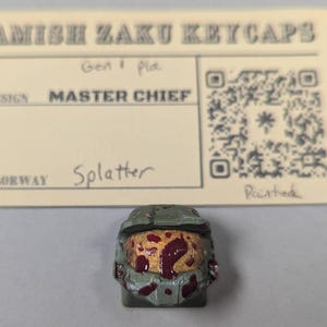 May include: A custom keycap featuring a Master Chief design with a splatter colorway. The keycap is painted with a green and red color scheme. The keycap is from Amish Zaku Keycaps.