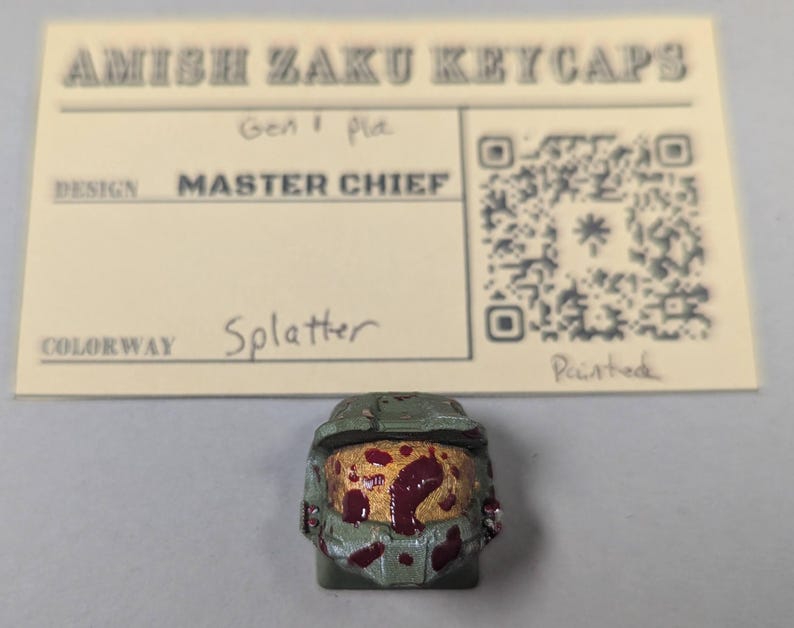 May include: A custom keycap featuring a Master Chief design with a splatter colorway. The keycap is painted with a green and red color scheme. The keycap is from Amish Zaku Keycaps.