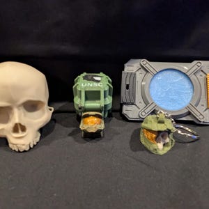 May include: A collection of toy items including a skull, a green and orange UNSC helmet, a gray and orange device with a blue center, and a green helmet keychain. The items are arranged on a dark surface.
