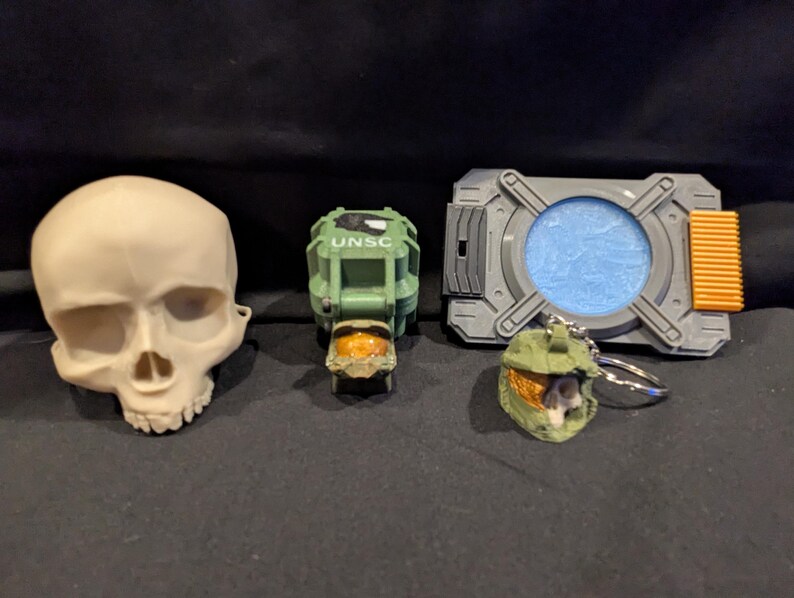 May include: A collection of toy items including a skull, a green and orange UNSC helmet, a gray and orange device with a blue center, and a green helmet keychain. The items are arranged on a dark surface.