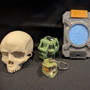 May include: A collection of 3D-printed items, including a skull, a green grenade with "UNSC" text, a gray and orange rectangular device, and a small green helmet keychain. The items are displayed on a dark surface.