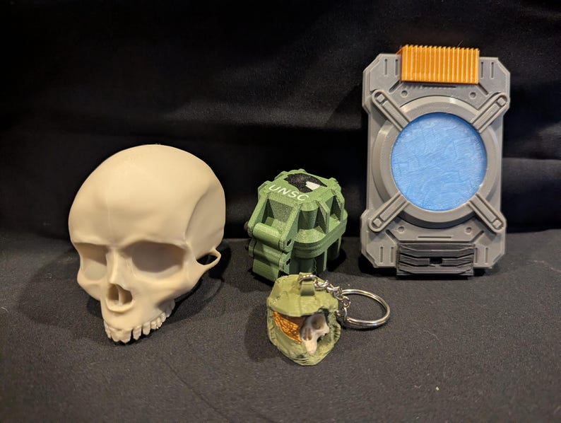 May include: A collection of 3D-printed items, including a skull, a green grenade with "UNSC" text, a gray and orange rectangular device, and a small green helmet keychain. The items are displayed on a dark surface.
