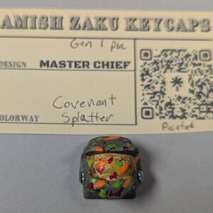 May include: A custom keycap featuring a Master Chief design with a Covenant Splatter colorway. The keycap is painted with a camouflage pattern of green, orange, and gold. The keycap is placed below a card that reads "Amish Zaku Keycaps".