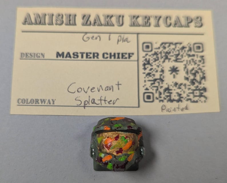 May include: A custom keycap featuring a Master Chief design with a Covenant Splatter colorway. The keycap is painted with a camouflage pattern of green, orange, and gold. The keycap is placed below a card that reads "Amish Zaku Keycaps".