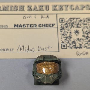 May include: A custom keycap in the shape of Master Chief's helmet, with a gold visor and green armor. The keycap is labeled "Midas Dust" and is made of resin. The card above the keycap reads "Amish Zaku Keycaps".