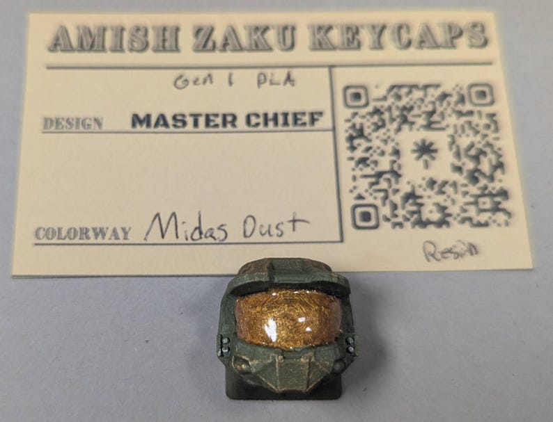 May include: A custom keycap in the shape of Master Chief's helmet, with a gold visor and green armor. The keycap is labeled "Midas Dust" and is made of resin. The card above the keycap reads "Amish Zaku Keycaps".