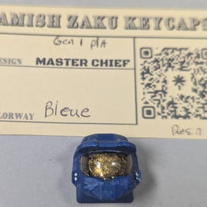 May include: A blue resin keycap shaped like a Master Chief helmet, with a gold visor. The keycap is part of the Amish Zaku Keycaps collection. The colorway is labeled as "Bleue" on a card behind the keycap.