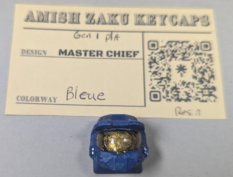May include: A blue resin keycap shaped like a Master Chief helmet, with a gold visor. The keycap is part of the Amish Zaku Keycaps collection. The colorway is labeled as "Bleue" on a card behind the keycap.