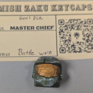 May include: A green and gold keycap in the shape of a Master Chief helmet, with a "Battle Worn" colorway. The keycap is placed in front of a beige card with the text "Amish Zaku Keycaps" and a QR code.