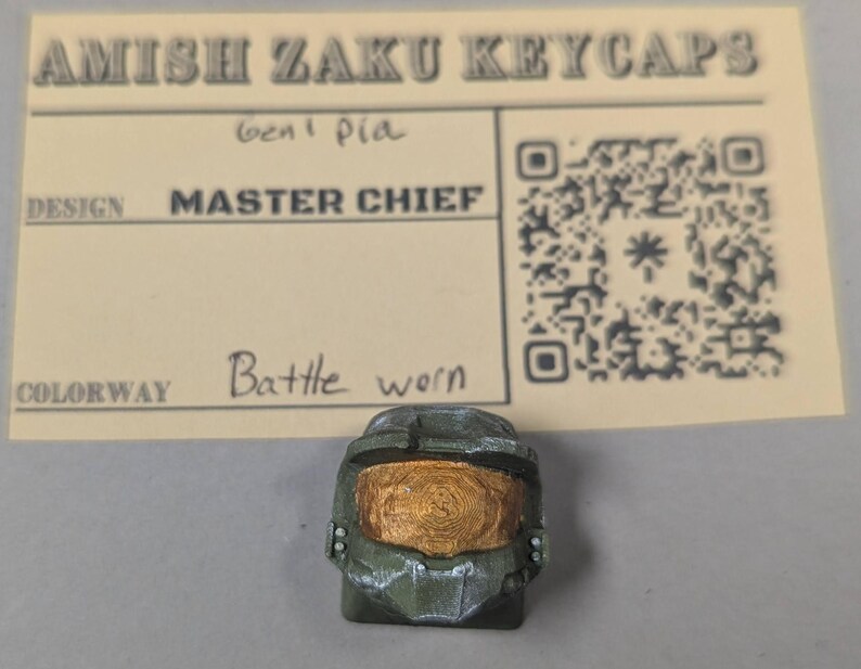 May include: A green and gold keycap in the shape of a Master Chief helmet, with a "Battle Worn" colorway. The keycap is placed in front of a beige card with the text "Amish Zaku Keycaps" and a QR code.