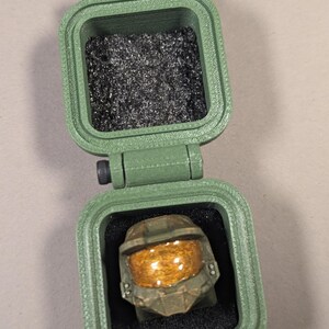 May include: A green, hinged box with a ring shaped like a helmet inside. The ring has a gold visor and is set in black foam. The top of the box is open, revealing a compartment filled with silver shavings.