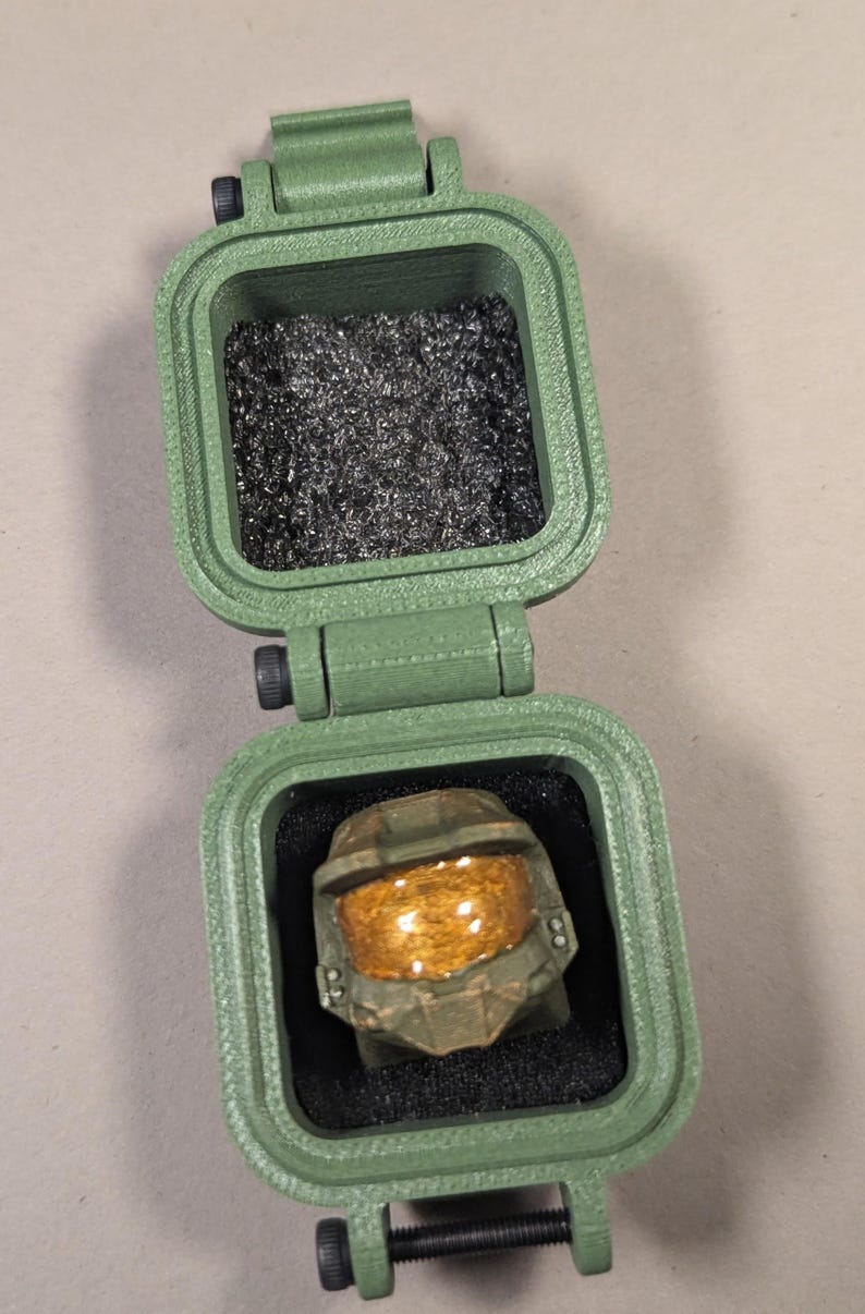 May include: A green, hinged box with a ring shaped like a helmet inside. The ring has a gold visor and is set in black foam. The top of the box is open, revealing a compartment filled with silver shavings.