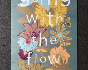 May include: A rectangular yoga mat featuring a floral design in shades of yellow, blue, purple, and orange. The words "going with the flow" are written in white across the mat. It is on a grey surface.