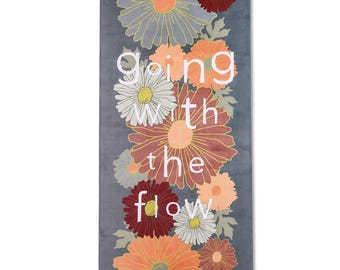 May include: A rectangular yoga mat with a floral design and the words "going with the flow." The mat features various flowers in shades of orange, red, white, and grey on a dark grey background. The brand name "flowology" is at the bottom.