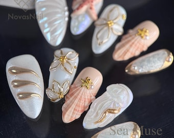 May include: A collection of handmade press-on nails with a sea-inspired theme. Designs include seashells, starfish, and floral accents in white, gold, and peach tones. The nails have a glossy finish and are arranged on a dark surface.