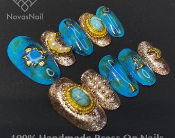 May include: A selection of handmade press-on nails in a turquoise and gold colour scheme. The nails are decorated with gold glitter, and feature designs including a sun, moon, and star. The text "NovasNail" and "100% Handmade Press On Nails" are visible.