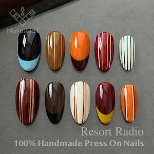 May include: Ten press-on nails featuring various designs including solid colours, vertical stripes, and colour-blocked patterns in shades of black, light blue, brown, orange, dark red, white, green, and yellow with gold accents. Text reads NovasNail Resort Radio.