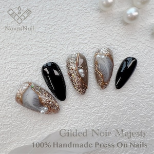 May include: Gilded Noir Majesty 100% Handmade Press On Nails.  Five press-on nails with a black, glitter, and gold design.  Two nails feature a white feather design with gold accents and pearl embellishments.