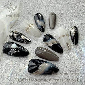 May include: Set of ten black and white press-on nails with different designs. The nails feature silver accents, glitter, and a pearl-like embellishment. The text "Aurora Noir" and "100% Handmade Press On Nails" is visible in the image.