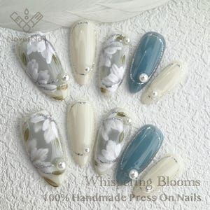 May include: Set of ten press-on nails with a floral design. The nails are in two colors: white and light blue. The nails have a glossy finish and are decorated with silver accents. The nails are arranged in a row on a white surface. The text "Whispering Blooms" and "100% Handmade Press On Nails" is visible in the image.