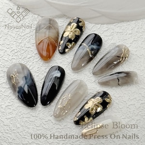 May include: Set of ten press-on nails with a black and gray marble design. Some nails feature gold accents and a floral design. The nails are labeled "Eclipse Bloom 100% Handmade Press On Nails".