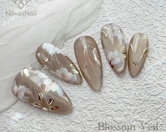 May include: Set of five beige press-on nails with delicate white floral designs and gold accents. The nails are arranged on a white background with the text "Blossom Veil 100% Handmade Press On Nails".