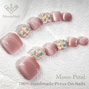 Puede incluir: Un conjunto de uñas postizas rosas con un acabado brillante. Algunas uñas presentan adornos iridiscentes con forma de gema. El texto "Moon Petal" y "100% Handmade Press On Nails" son visibles.